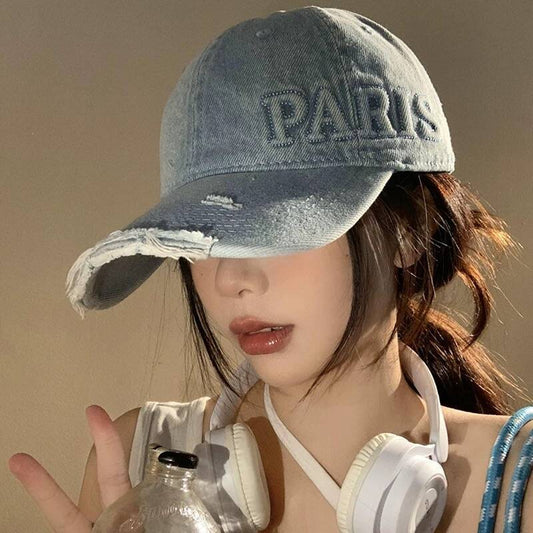 Female model wearing a washed denim baseball cap with distressed holes and "PARIS" embroidered on the front, paired with white headphones around the neck.